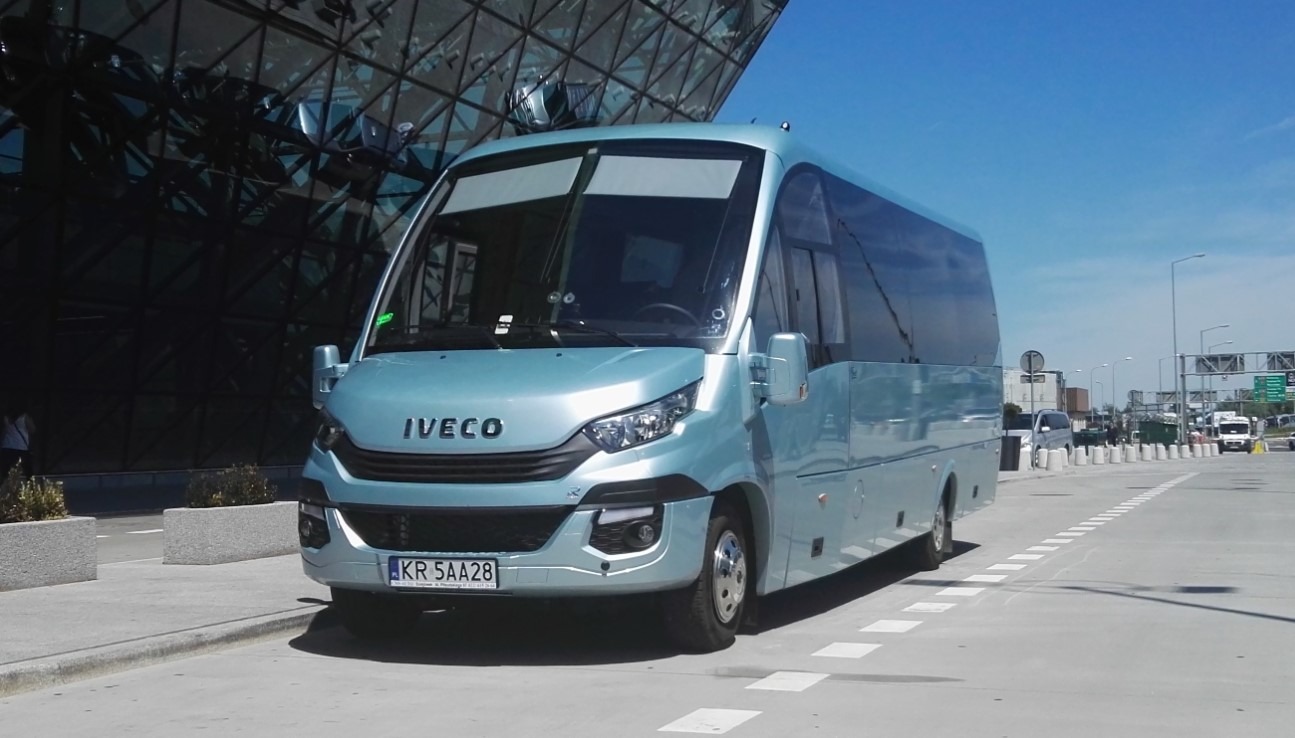 Niebieski bus Iveco z numerem rejestracyjnym KR 5AA28 na tle nowoczesnego budynku lotniska w słoneczny dzień.