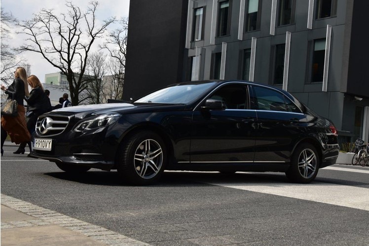 Czarna limuzyna Mercedes-Benz zaparkowana na pasach dla pieszych przed nowoczesnym budynkiem biurowym, w tle przechodzący ludzie.