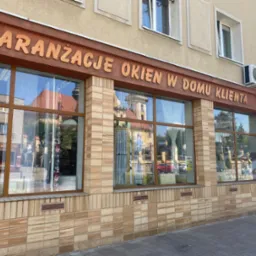 Fasada budynku z dużymi oknami wystawowymi i napisem 'ARANŻACJE OKIEN W DOMU KLIENTA', w szybach okien odbijają się budynki i niebo.