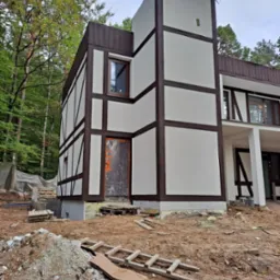 Dom w stylu szachulcowym w trakcie ocieplania elewacji, widoczna folia ochronna na drzwiach wejściowych, rusztowania leżące na ziemi.