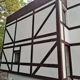 Fragment budynku w stylu szachulcowym z brązowymi belkami i jasną elewacją, widoczne fundamenty w trakcie prac wykończeniowych, otoczenie leśne.