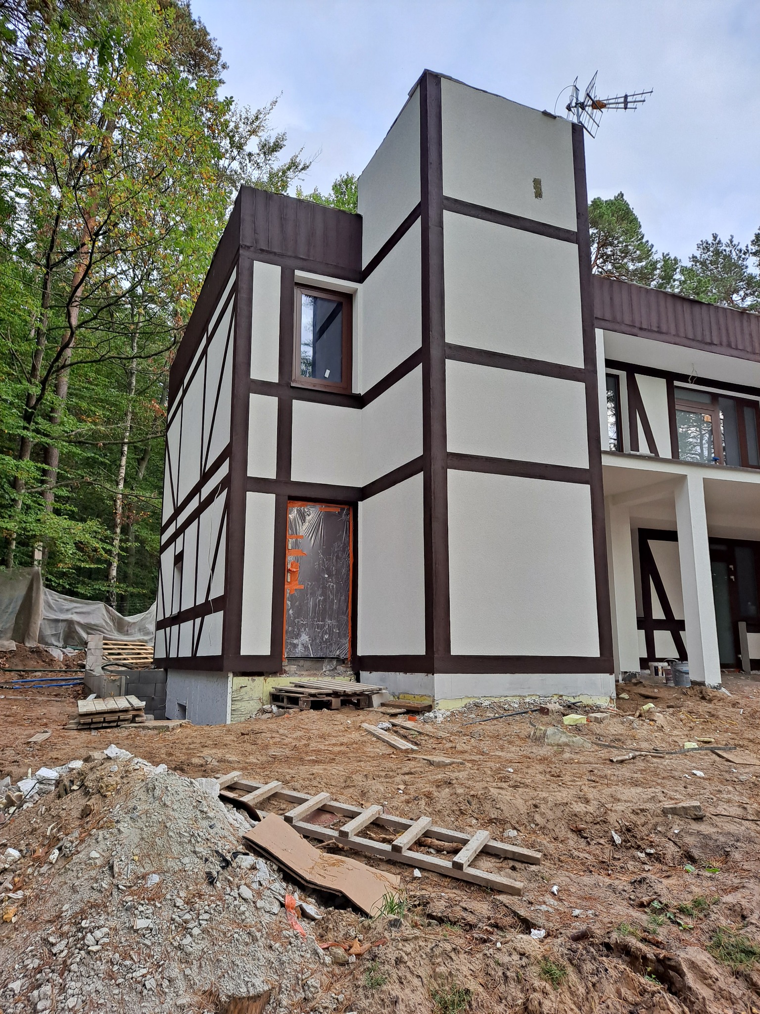 Dom w stylu szachulcowym w trakcie ocieplania elewacji, widoczna folia ochronna na drzwiach wejściowych, rusztowania leżące na ziemi.