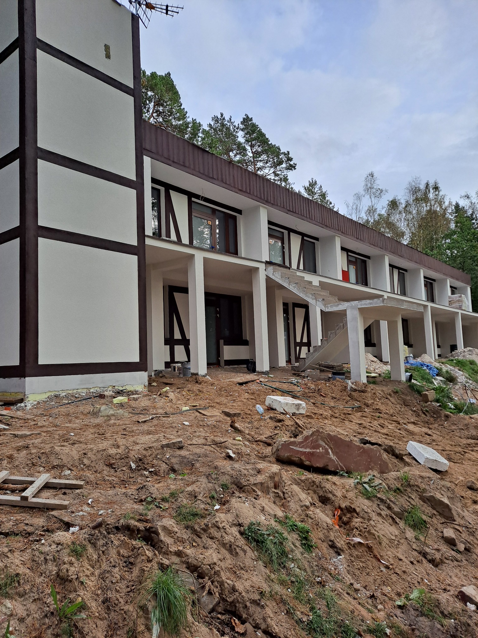 Budynek w trakcie prac elewacyjnych, widoczne fragmenty ocieplenia przy fundamentach, świeżo pomalowana elewacja z elementami dekoracyjnymi imitującymi konstrukcję szkieletową, teren wokół budynku...