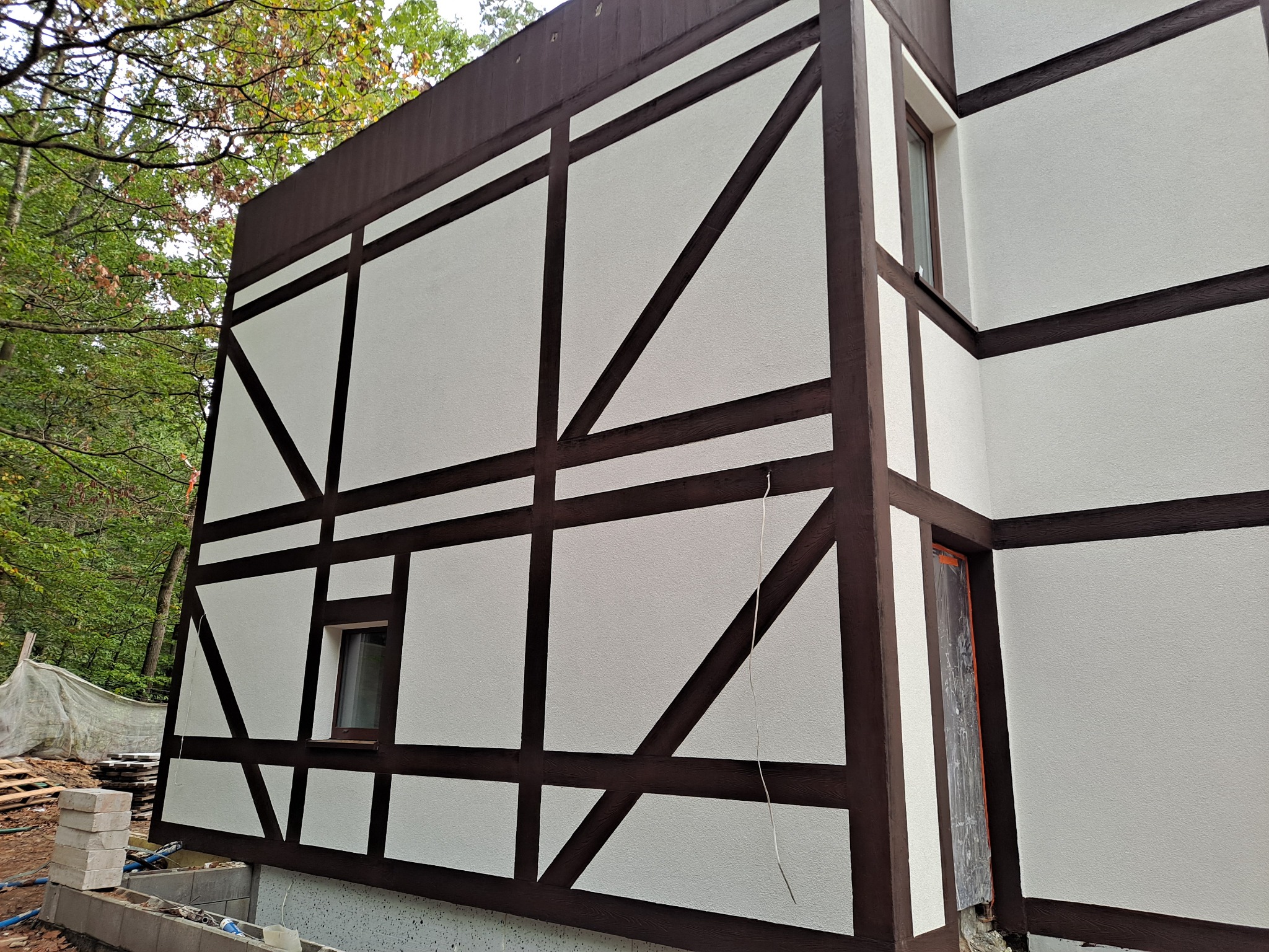 Fragment budynku w stylu szachulcowym z brązowymi belkami i jasną elewacją, widoczne fundamenty w trakcie prac wykończeniowych, otoczenie leśne.