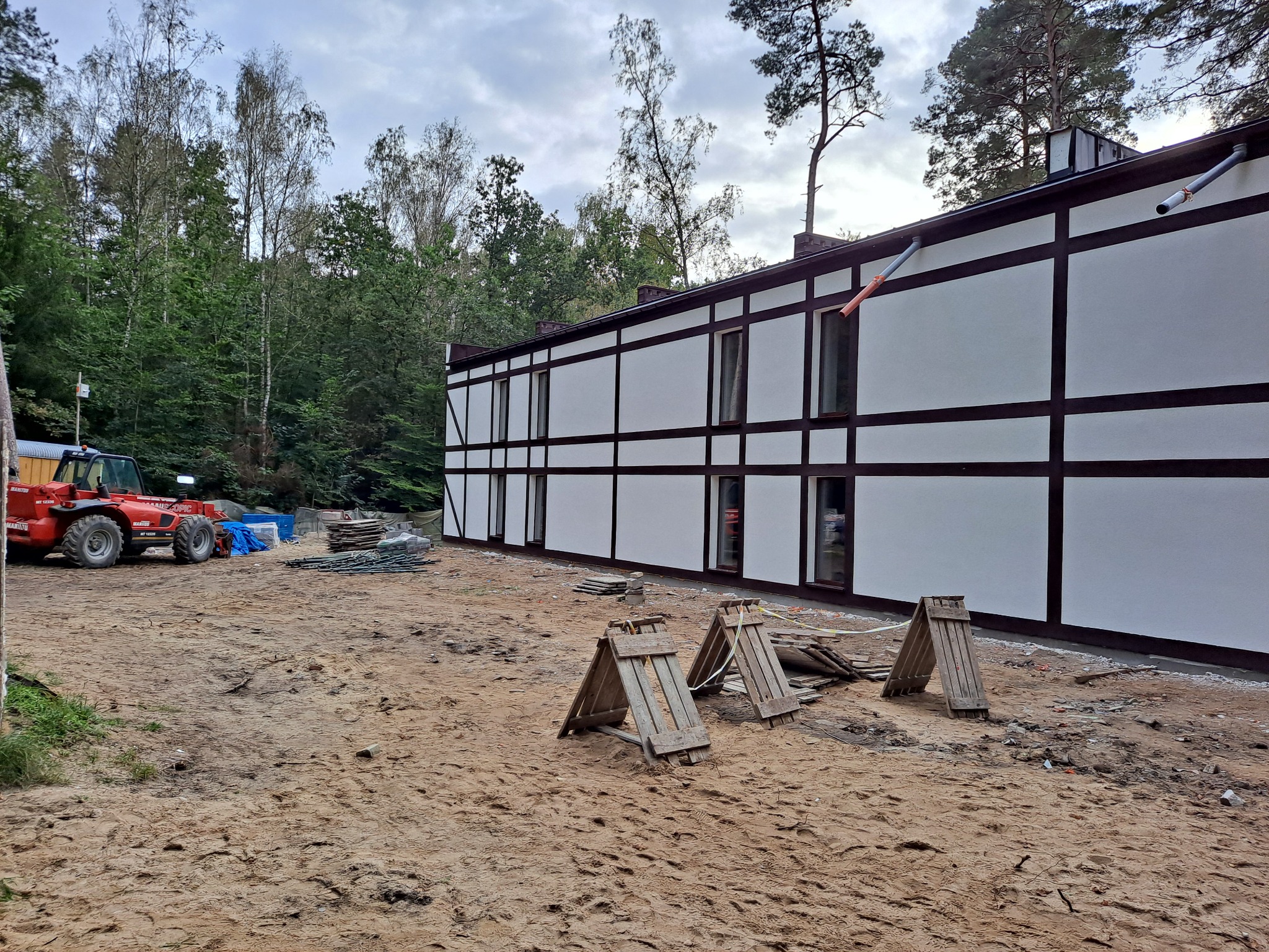 Biała elewacja budynku w stylu szachownicy z brązowymi belkami, widoczny czerwony wózek widłowy Manitou, drewniane kozły i materiały budowlane na piaszczystym terenie.