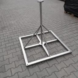 Metalowa konstrukcja wsporcza z kwadratową podstawą i centralnym słupkiem, widoczna na szarym bruku, prawdopodobnie element montażowy do instalacji zewnętrznej.