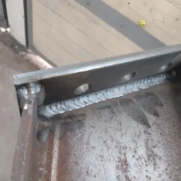 Spoina spawalnicza łącząca dwie metalowe płyty, widoczne ślady korozji na powierzchni metalu i otwory montażowe.