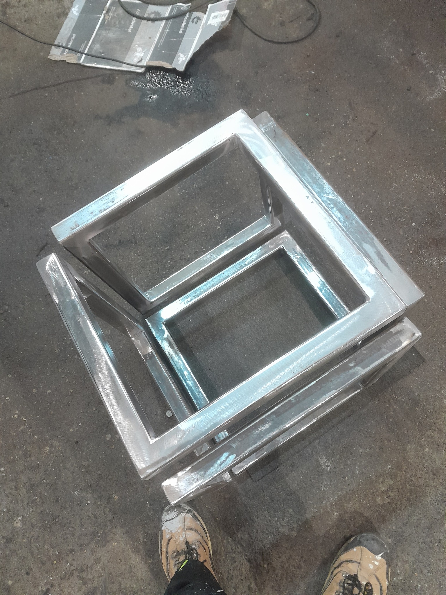 Metalowe ramy o kwadratowym przekroju, ułożone jedna na drugiej, widoczne ślady obróbki, surowy beton na podłodze, fragment buta w rogu kadru.