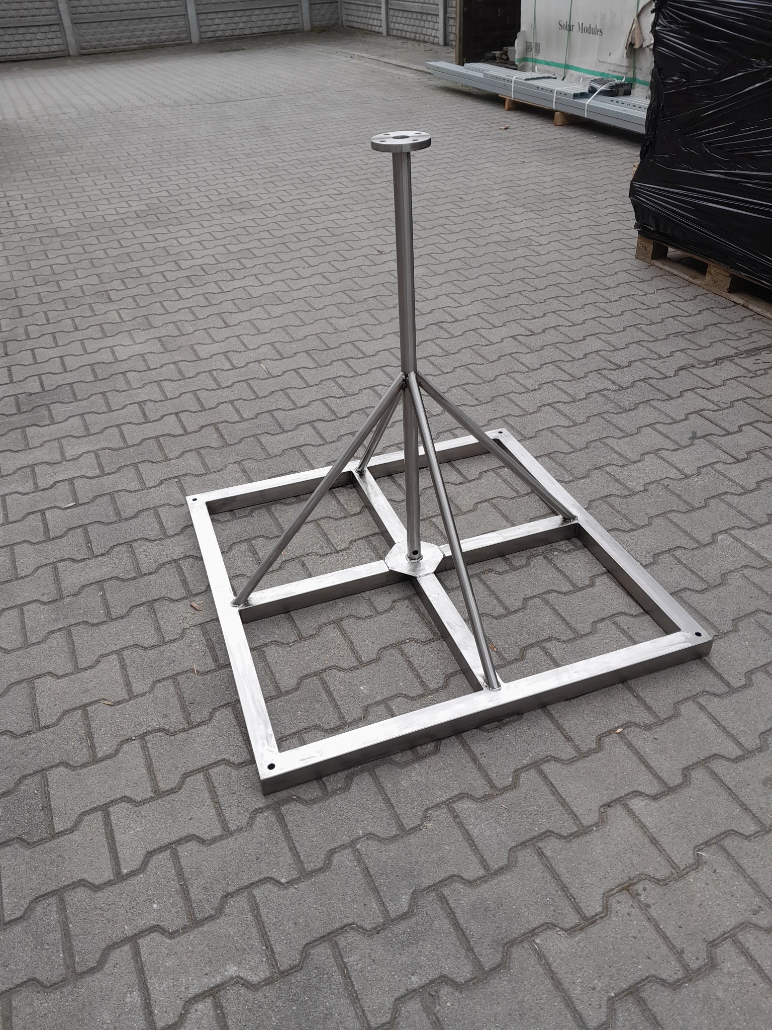 Metalowa konstrukcja wsporcza z kwadratową podstawą i centralnym słupkiem, widoczna na szarym bruku, prawdopodobnie element montażowy do instalacji zewnętrznej.