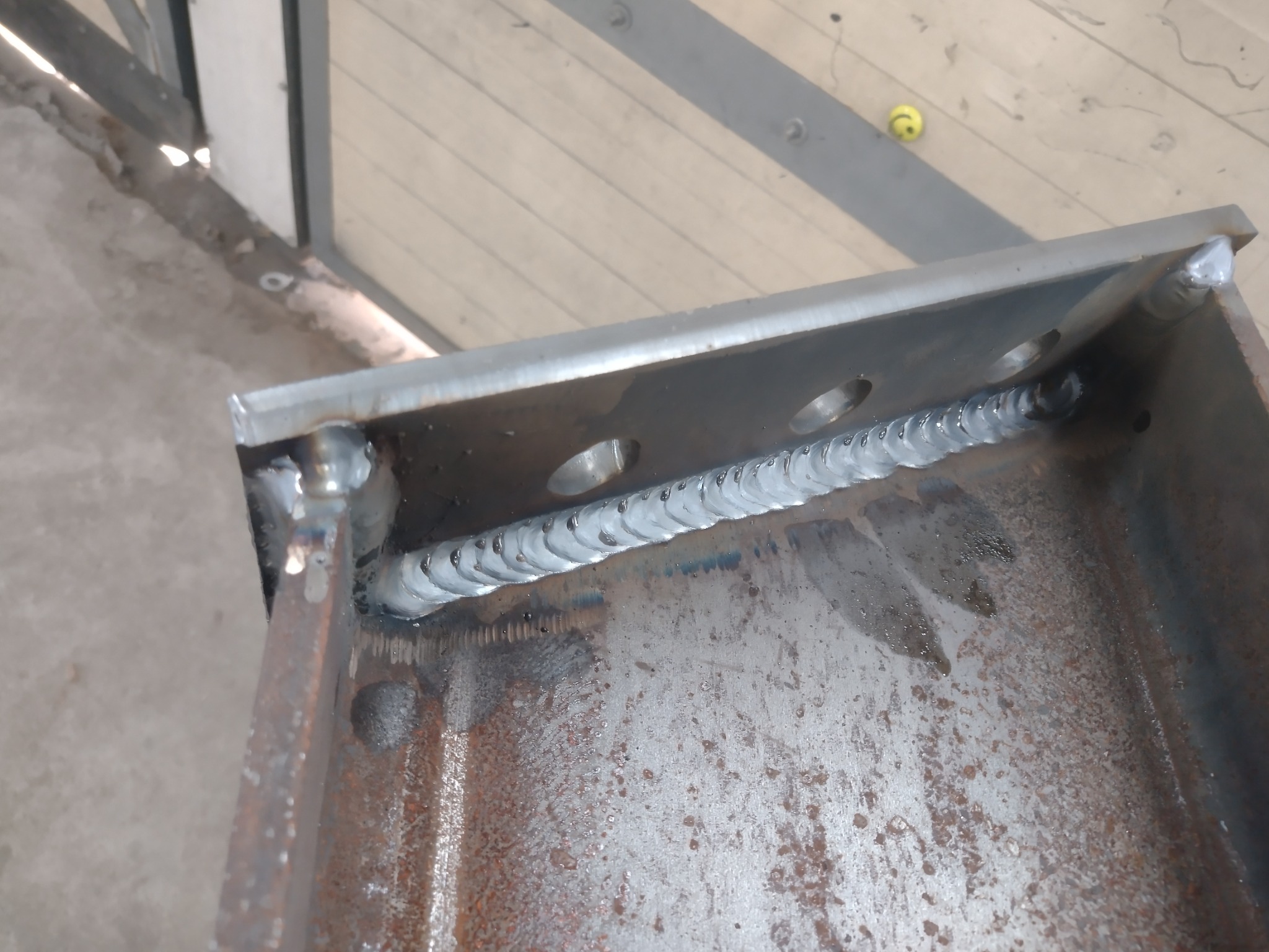 Spoina spawalnicza łącząca dwie metalowe płyty, widoczne ślady korozji na powierzchni metalu i otwory montażowe.