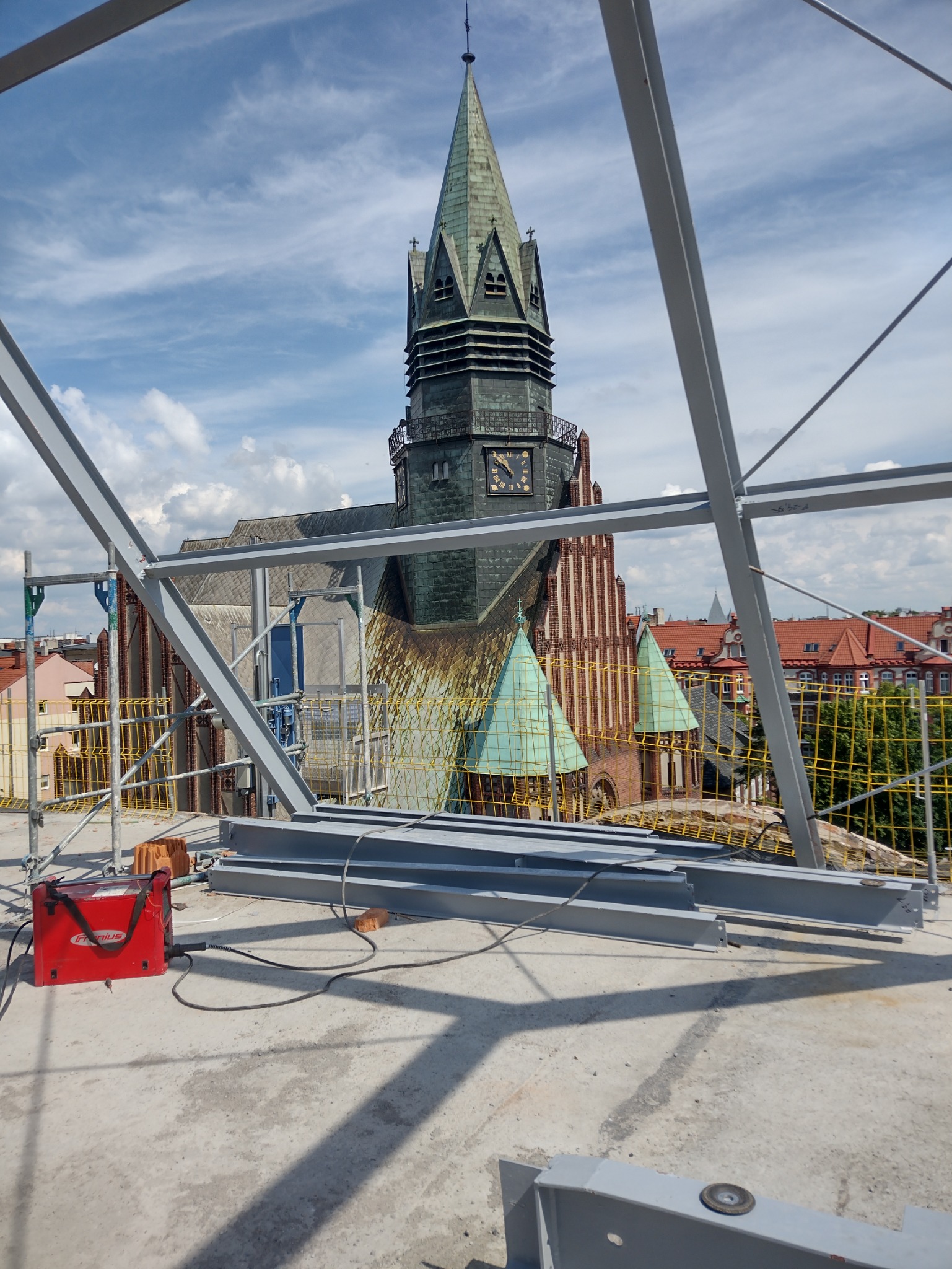 W trakcie budowy konstrukcji stalowej w Poznaniu, z widokiem na wieżę kościoła. Na pierwszym planie leżą elementy metalowe i czerwona spawarka.