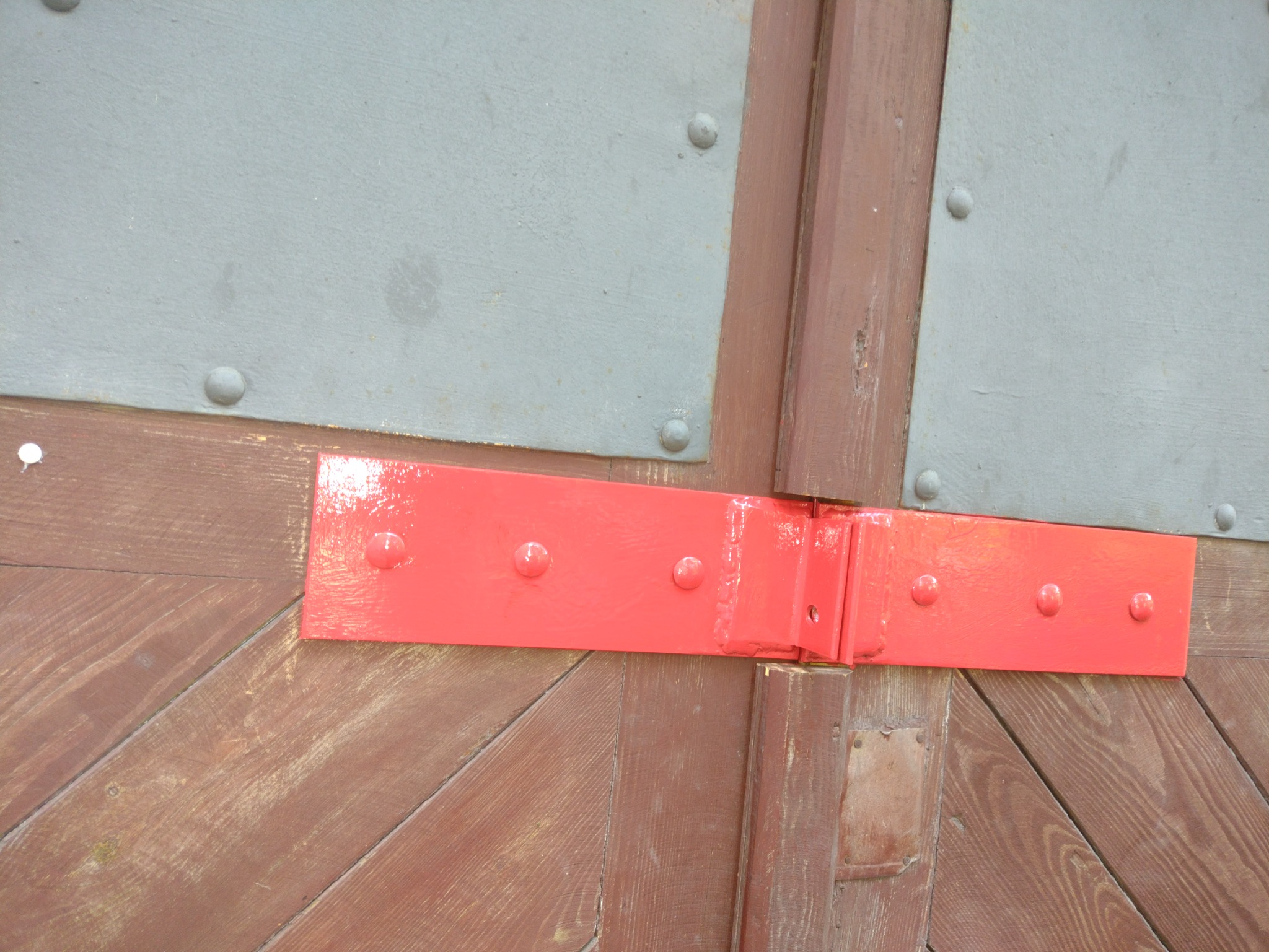 Fragment drewnianej bramy z metalowymi elementami i czerwonym, pomalowanym na wysoki połysk zawiasem z nitami.