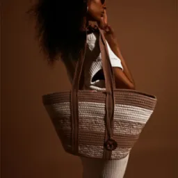 Portret kobiety w okularach, z torebką na ramieniu na jednolitym, brązowym tle. Ujęcie z profilu, z delikatnym oświetleniem podkreślającym detale torebki.