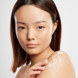 Kamil Słupecki FotograVita - Portret kobiety z nałożonym peelingiem na ramieniu, dłoń delikatnie dotyka skóry. Jasne tło, naturalne światło, widoczna struktura produktu kosmetycznego.