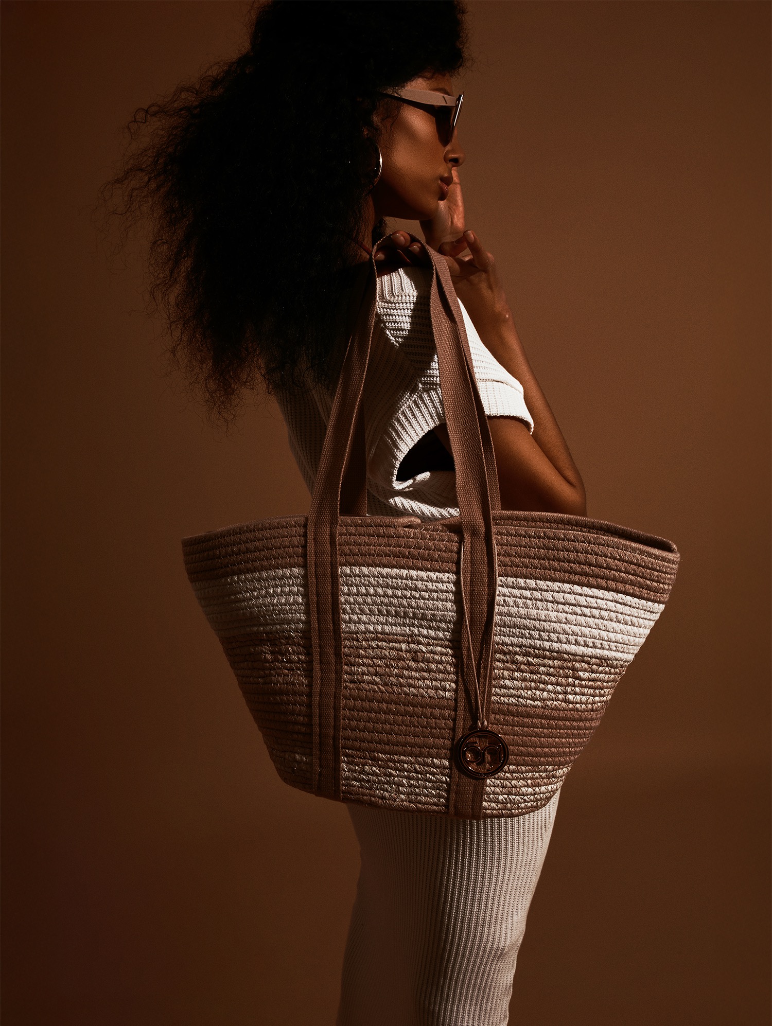 Portret kobiety w okularach, z torebką na ramieniu na jednolitym, brązowym tle. Ujęcie z profilu, z delikatnym oświetleniem podkreślającym detale torebki.