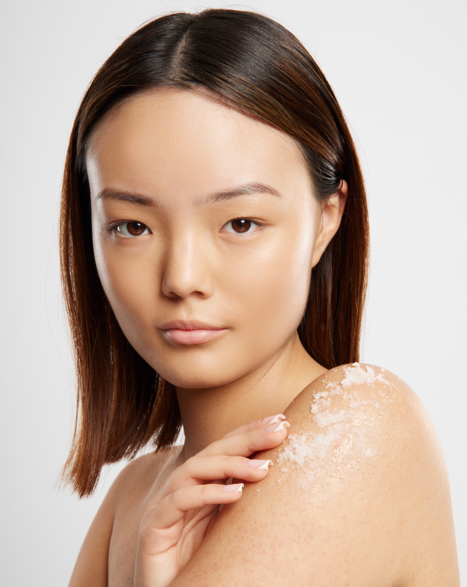 Portret kobiety z nałożonym peelingiem na ramieniu, dłoń delikatnie dotyka skóry. Jasne tło, naturalne światło, widoczna struktura produktu kosmetycznego.