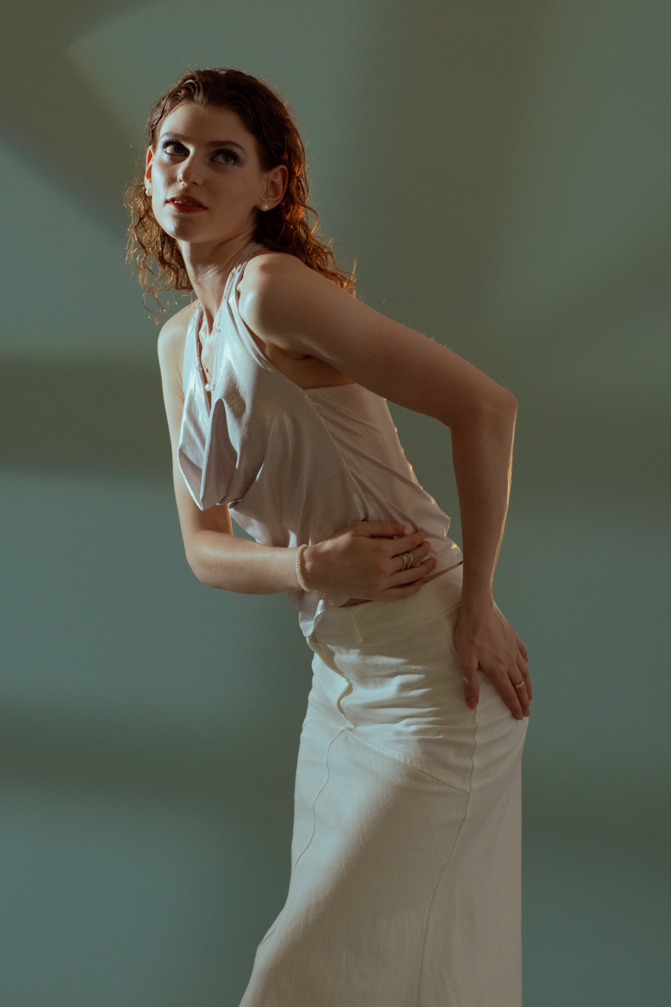 Portret kobiety w jasnej sukience, z delikatnym makijażem i mokrymi włosami, pozującej na rozmytym, zielonkawym tle w studiu fotograficznym. Ujęcie od dołu.
