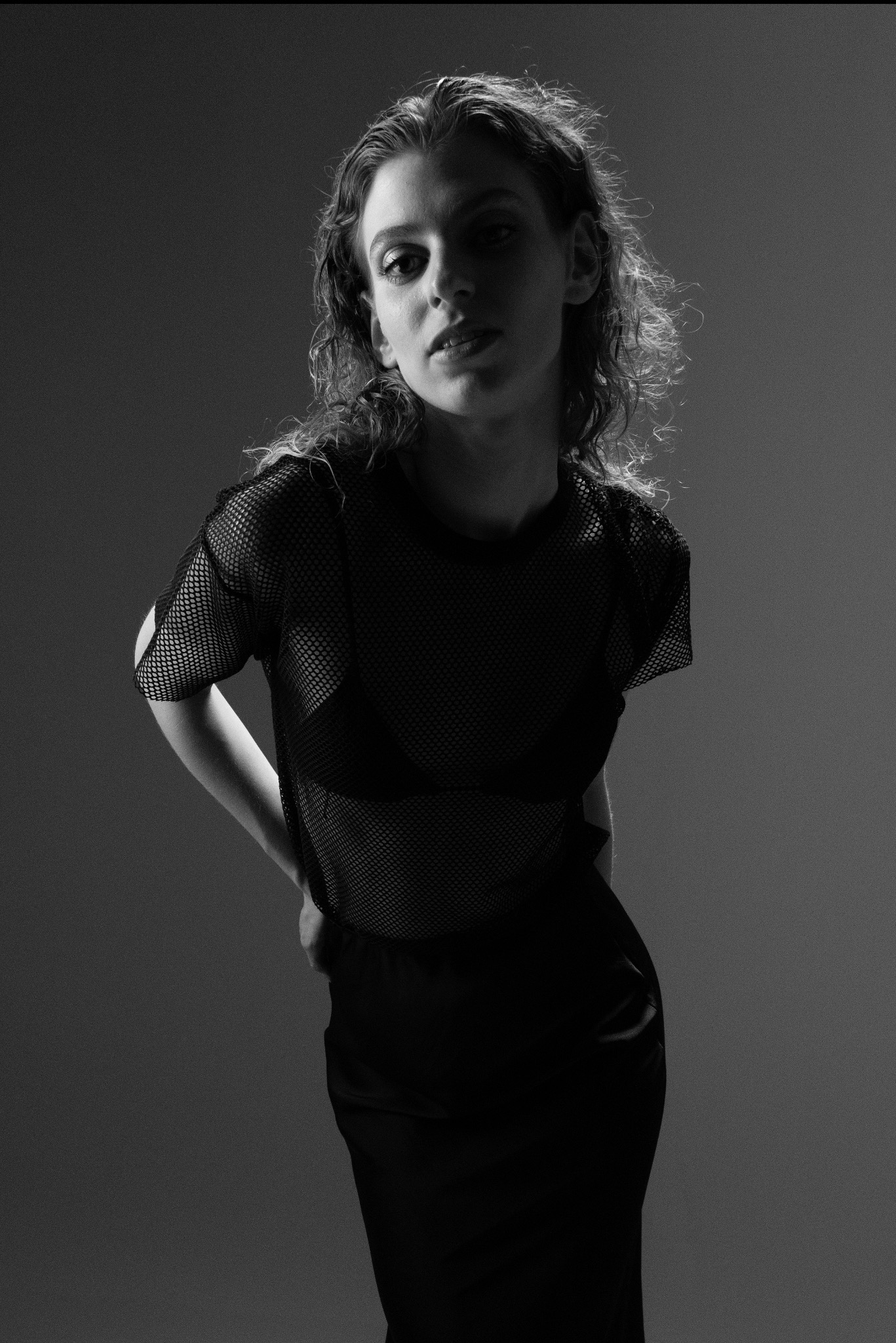Czarno-białe, studyjne zdjęcie kobiety w siatkowej bluzce i spódnicy, z rękami na biodrach, w półmroku, z delikatnym oświetleniem podkreślającym rysy twarzy.