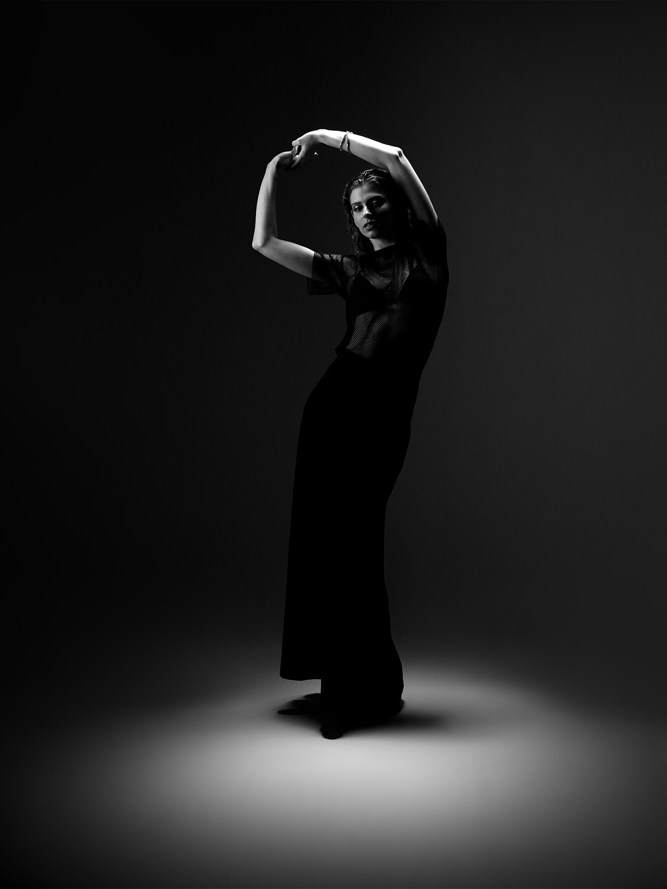 Czarno-białe, pełne ujęcie kobiety w ciemnej, prześwitującej bluzce i długiej spódnicy, pozującej z uniesionymi rękami, oświetlonej punktowo od dołu na ciemnym tle.
