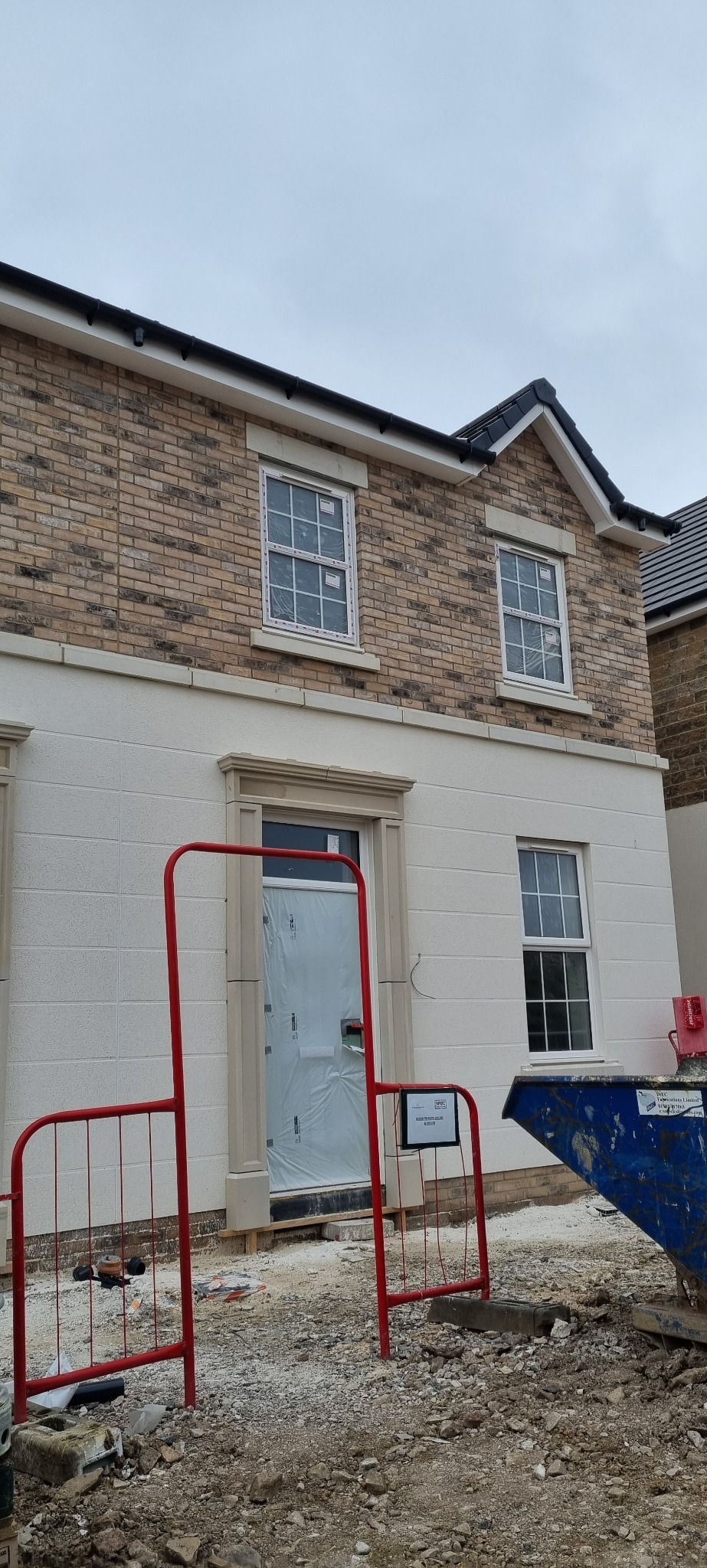 Elewacja domu jednorodzinnego w trakcie prac wykończeniowych, widoczne rusztowanie, kontener na gruz i zabezpieczone folią okna oraz drzwi.