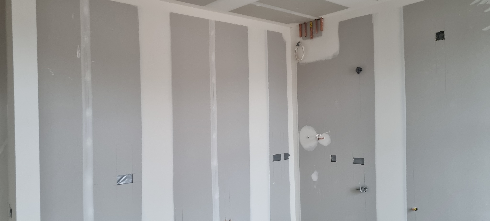 Wnętrze w trakcie prac wykończeniowych: widoczne płyty gipsowo-kartonowe na ścianach i suficie, z instalacjami elektrycznymi i wodnymi przygotowanymi do dalszych prac.