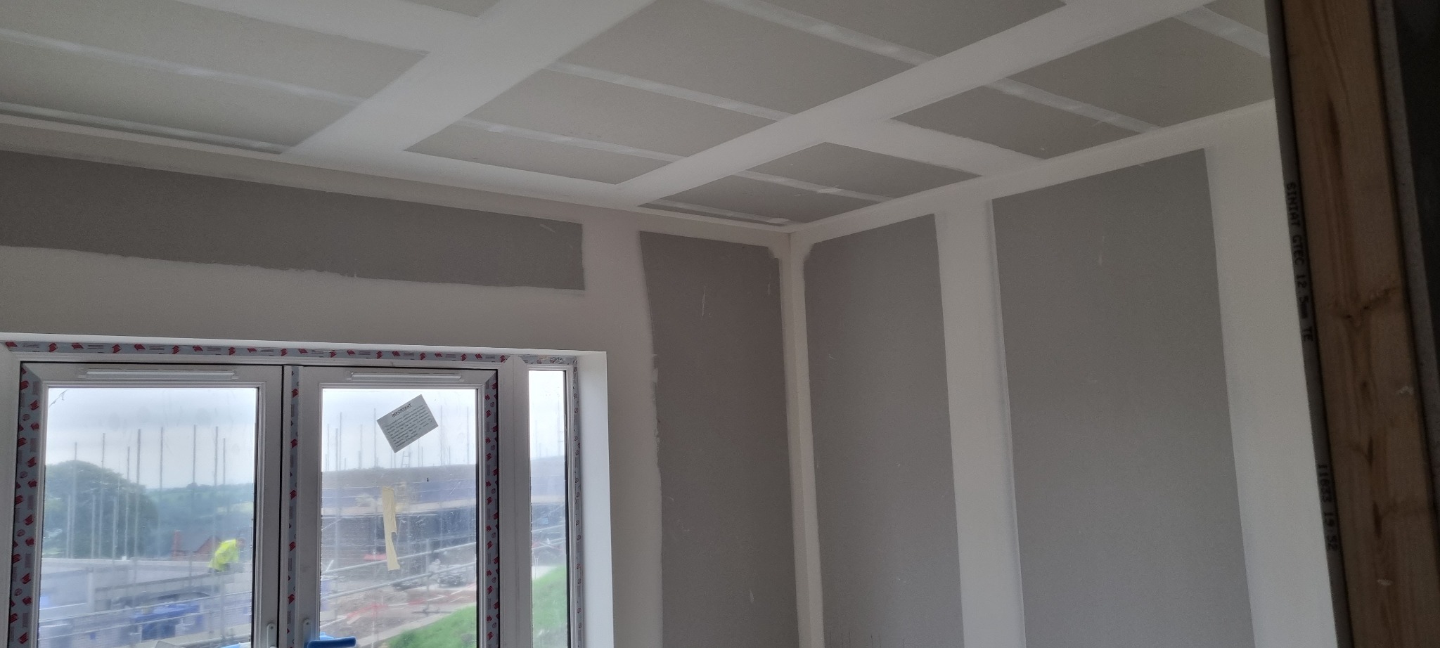 Montaż płyt kartonowo-gipsowych na ścianach i suficie, widoczne łączenia płyt i wstępne szpachlowanie, okno z widokiem na budowę