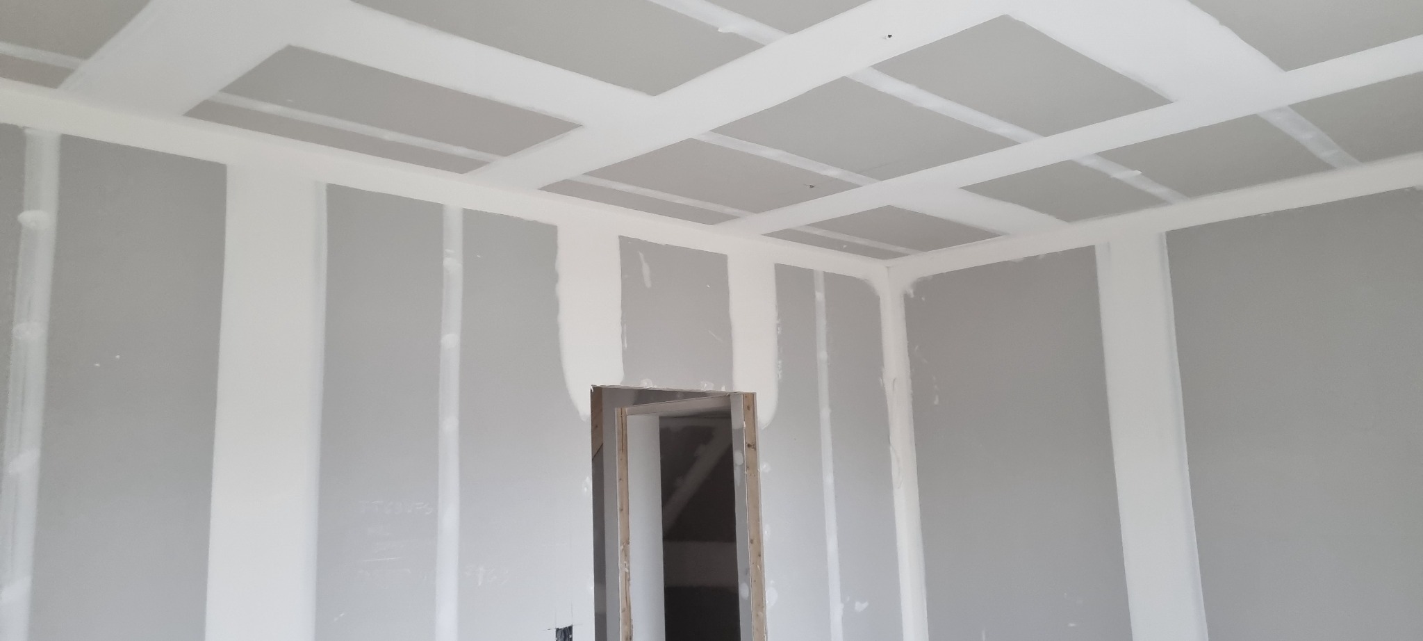 Wnętrze pokoju w trakcie montażu płyt gipsowo-kartonowych na ścianach i suficie, widoczne łączenia płyt wzmocnione taśmą. Otwór drzwiowy bez ościeżnicy.