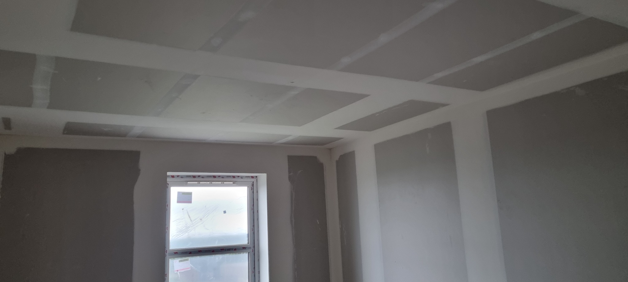 Wnętrze w trakcie prac wykończeniowych: ściany z fragmentami nałożonej gładzi szpachlowej, sufit z widocznymi łączeniami płyt kartonowo-gipsowych przed szlifowaniem, okno zabezpieczone folią.