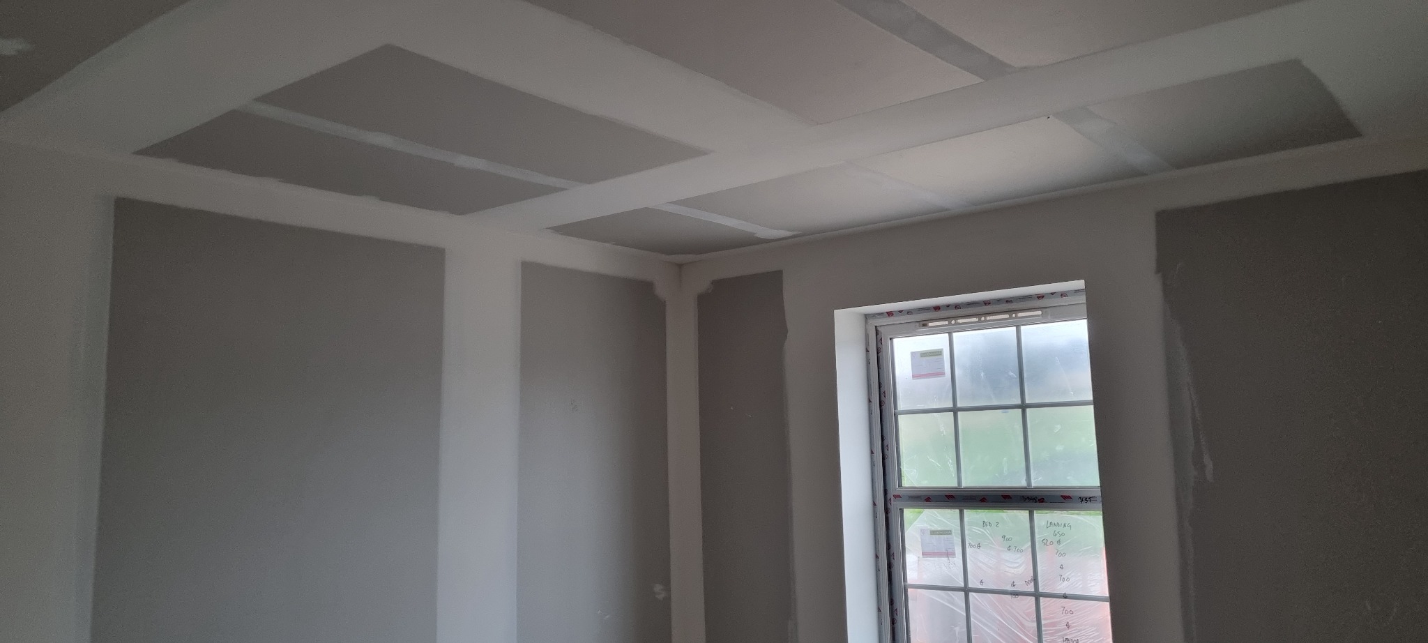 Pokój w trakcie remontu, ściany i sufit pokryte szarą warstwą, widoczne płyty na suficie z wyznaczonymi prostokątami, okno zabezpieczone folią.