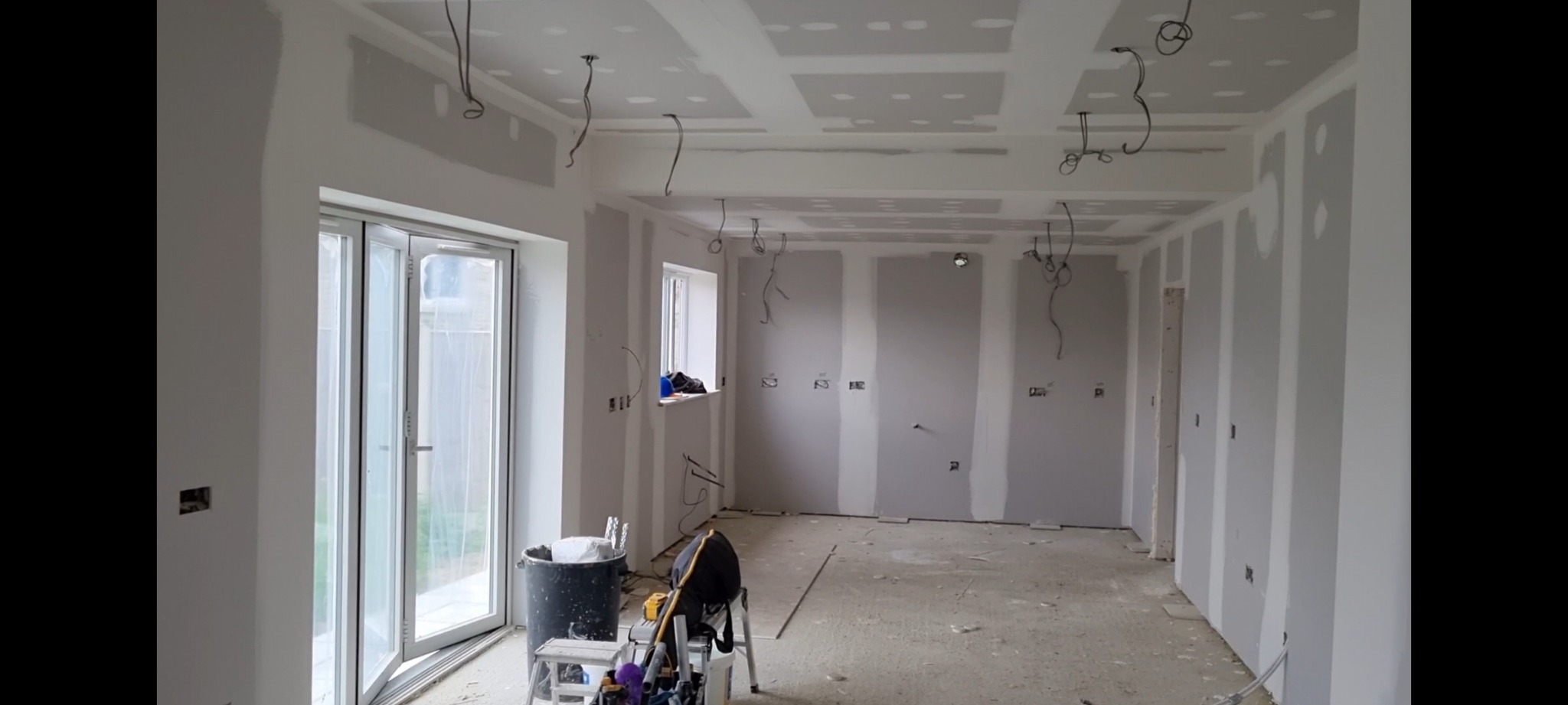 Wnętrze w trakcie remontu: ściany obłożone płytami gipsowo-kartonowymi, widoczne łączenia do wypełnienia, wystające kable elektryczne, drabina malarska i wiadro z narzędziami.