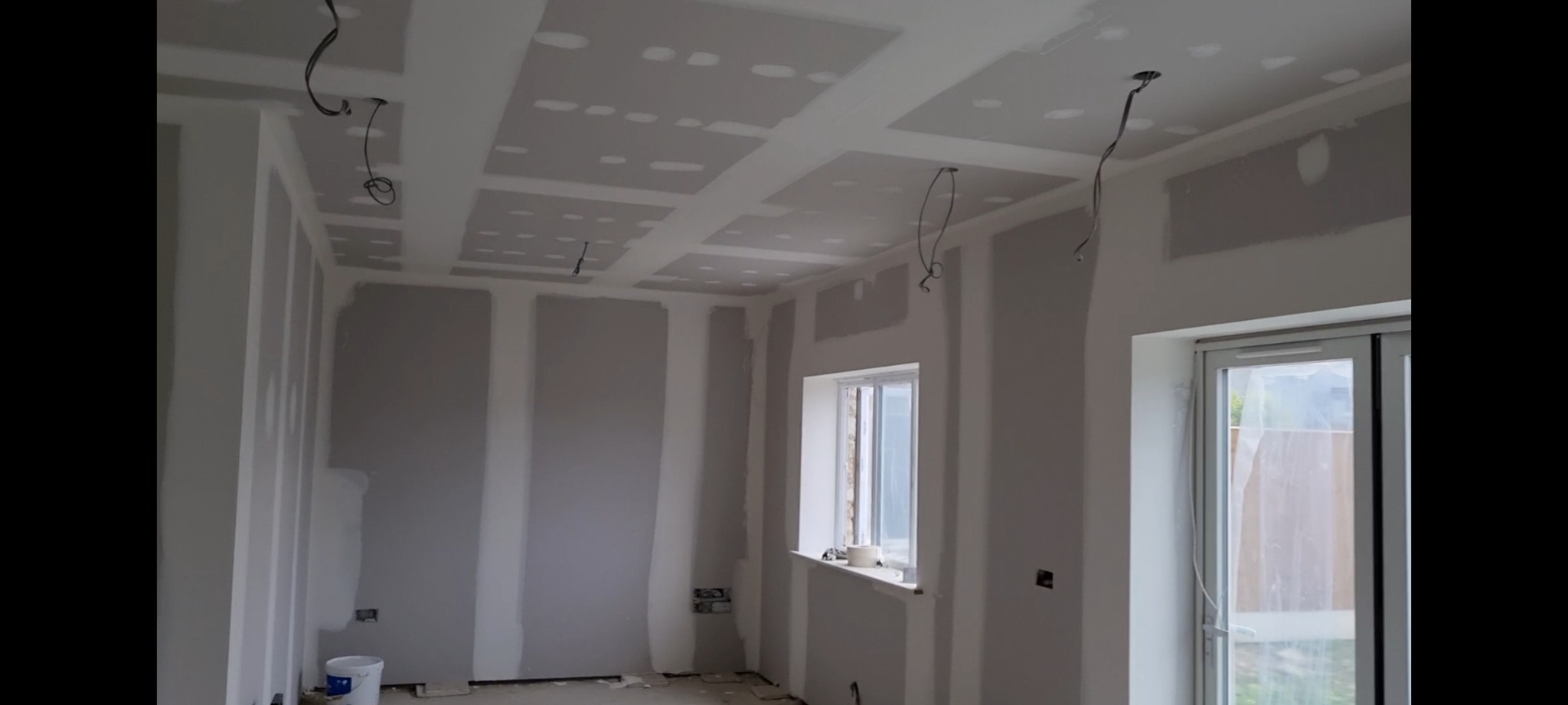 Pokój w trakcie remontu: ściany i sufit z płyt kartonowo-gipsowych, widoczne łączenia i otwory po szpachlowaniu, wystające kable elektryczne, białe okno z parapetem, wiadro farby na podłodze.