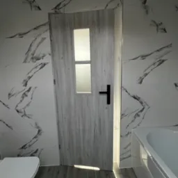 Wykończona łazienka z białymi płytkami imitującymi marmur, jasnymi panelami podłogowymi i drewnopodobnymi drzwiami z czarną klamką i przeszkleniami, widoczna toaleta i wanna.