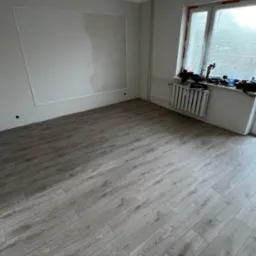 Jasne panele podłogowe ułożone w pokoju w Słupsku, ściana z dekoracyjną listwą, grzejnik pod oknem, surowa żarówka.