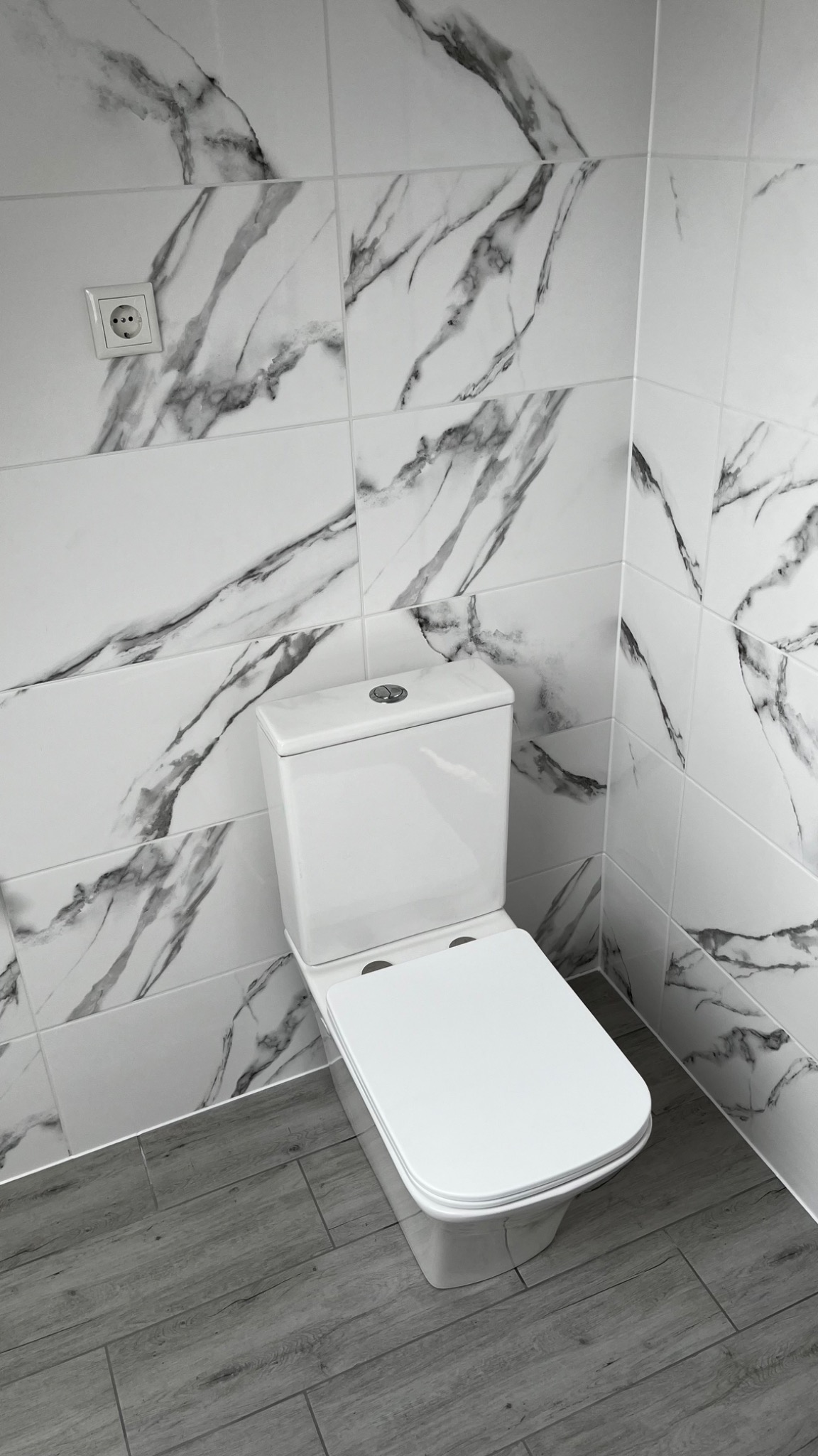 Biała toaleta kompaktowa z deską wolnoopadającą na tle ściany wyłożonej białymi płytkami imitującymi marmur i podłogi z paneli w odcieniach szarości; widoczny kontakt elektryczny.
