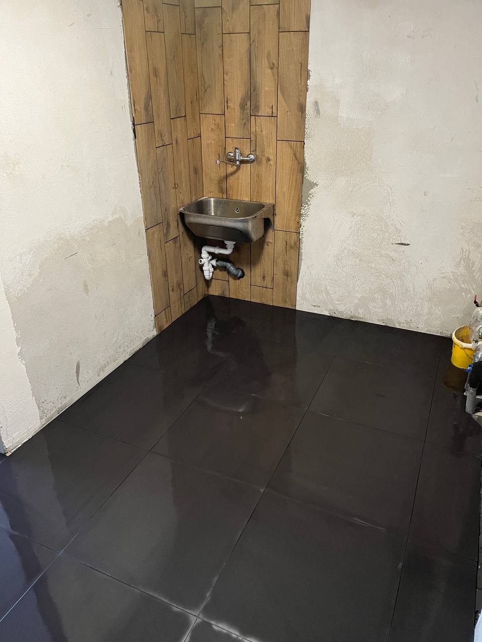 Łazienka w trakcie prac: ułożone ciemne, błyszczące płytki podłogowe, fragment ściany z drewnopodobnymi płytkami, zamontowana metalowa umywalka, ściana częściowo przygotowana pod malowanie.