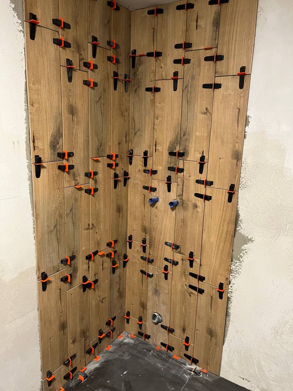 Ściana w trakcie układania płytek imitujących drewno, z użyciem pomarańczowych klipsów poziomujących, widoczne wystające rury instalacji wodnej i kanalizacyjnej.