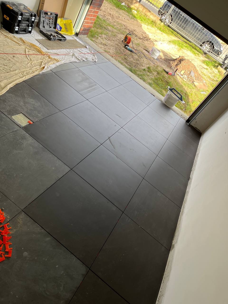 Ułożone, ciemnoszare płytki podłogowe w garażu, z pomarańczowymi krzyżykami dystansowymi, widoczne narzędzia i materiały budowlane w tle.