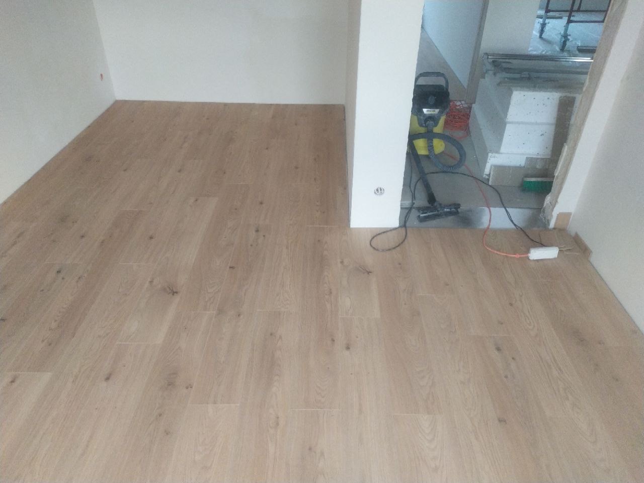 Układanie paneli podłogowych w jasnym odcieniu drewna, widoczny odkurzacz przemysłowy w przejściu do kolejnego pomieszczenia w trakcie sprzątania po montażu.