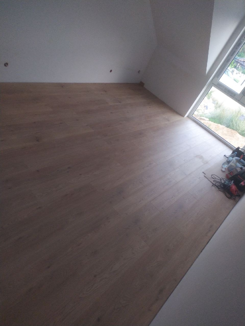 Wykończony pokój na poddaszu z nową, jasną podłogą drewnopodobną ułożoną w kierunku okna, narzędzia i materiały do wykończenia w rogu.