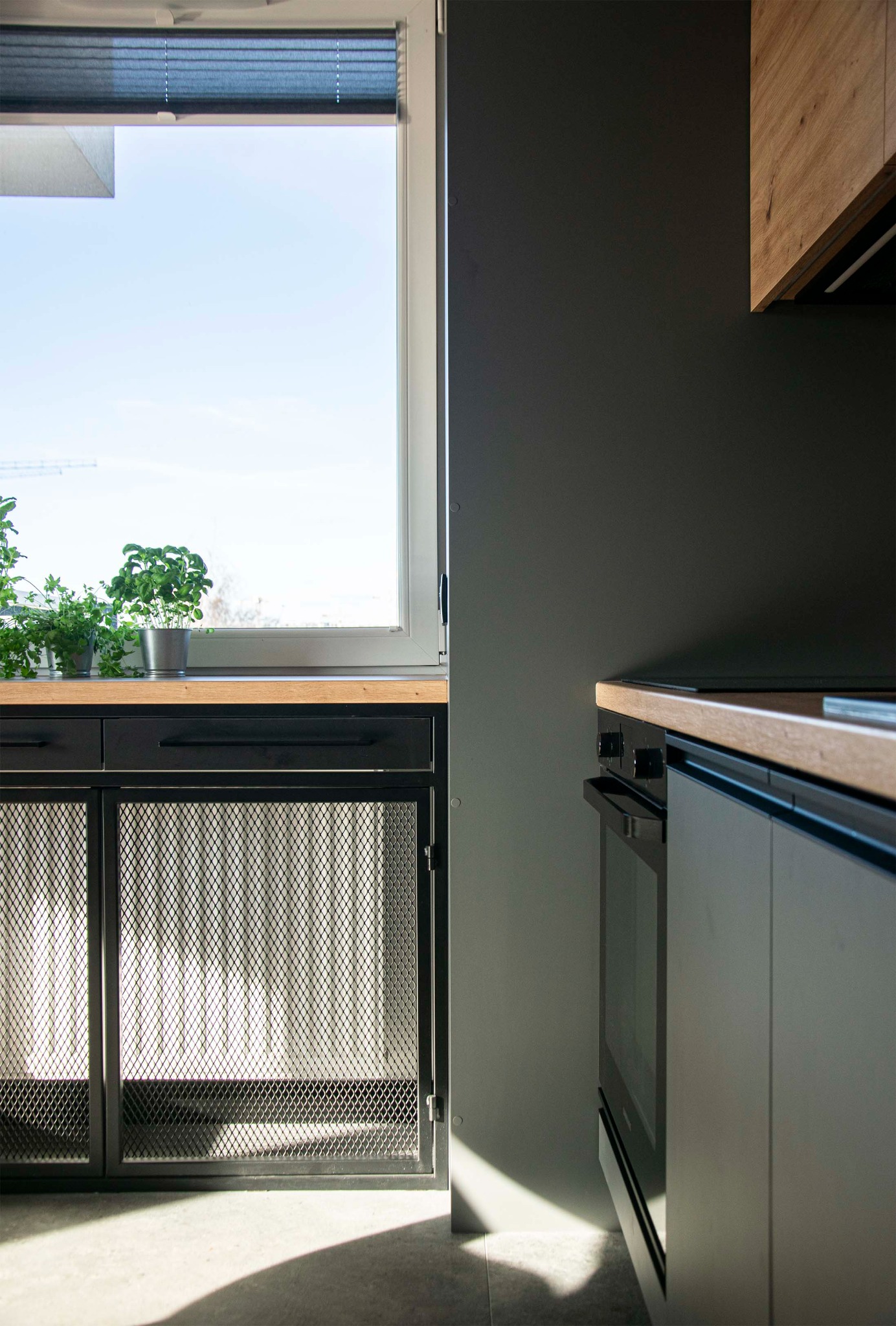 Fragment nowoczesnej kuchni z szarymi szafkami, drewnianym blatem, metalową siatką na frontach szafek dolnych i ziołami w doniczkach na parapecie okna.