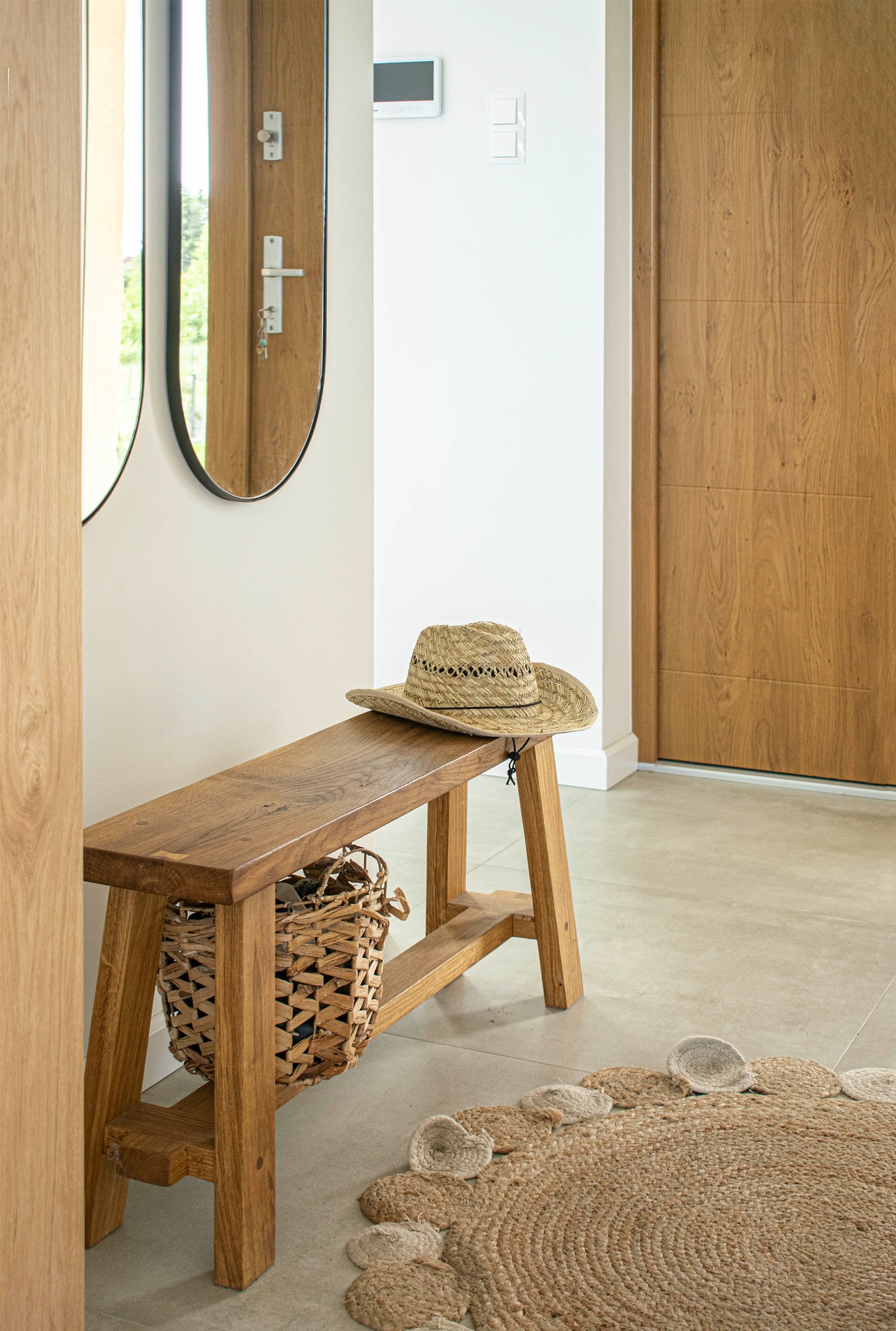 Jasne wnętrze przedpokoju z drewnianą ławką, na której leży słomkowy kapelusz, pod ławką wiklinowy kosz, na podłodze okrągły dywanik z jutowego sznurka.