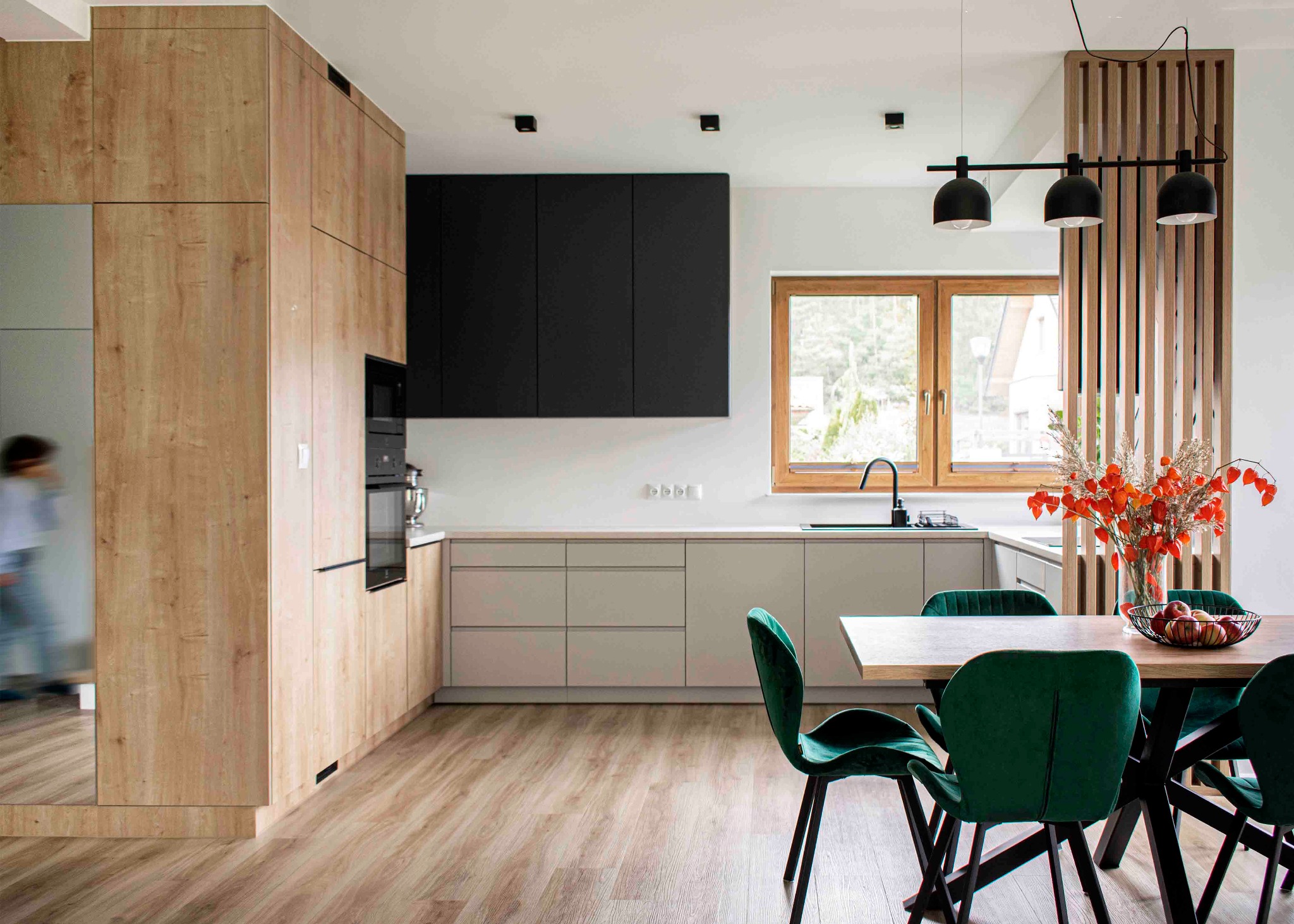 Nowoczesna kuchnia z drewnianymi i czarnymi szafkami, szarym blatem, drewnianym stołem z zielonymi krzesłami, oświetlona trzema czarnymi lampami wiszącymi i z widokiem na zewnątrz przez okno.