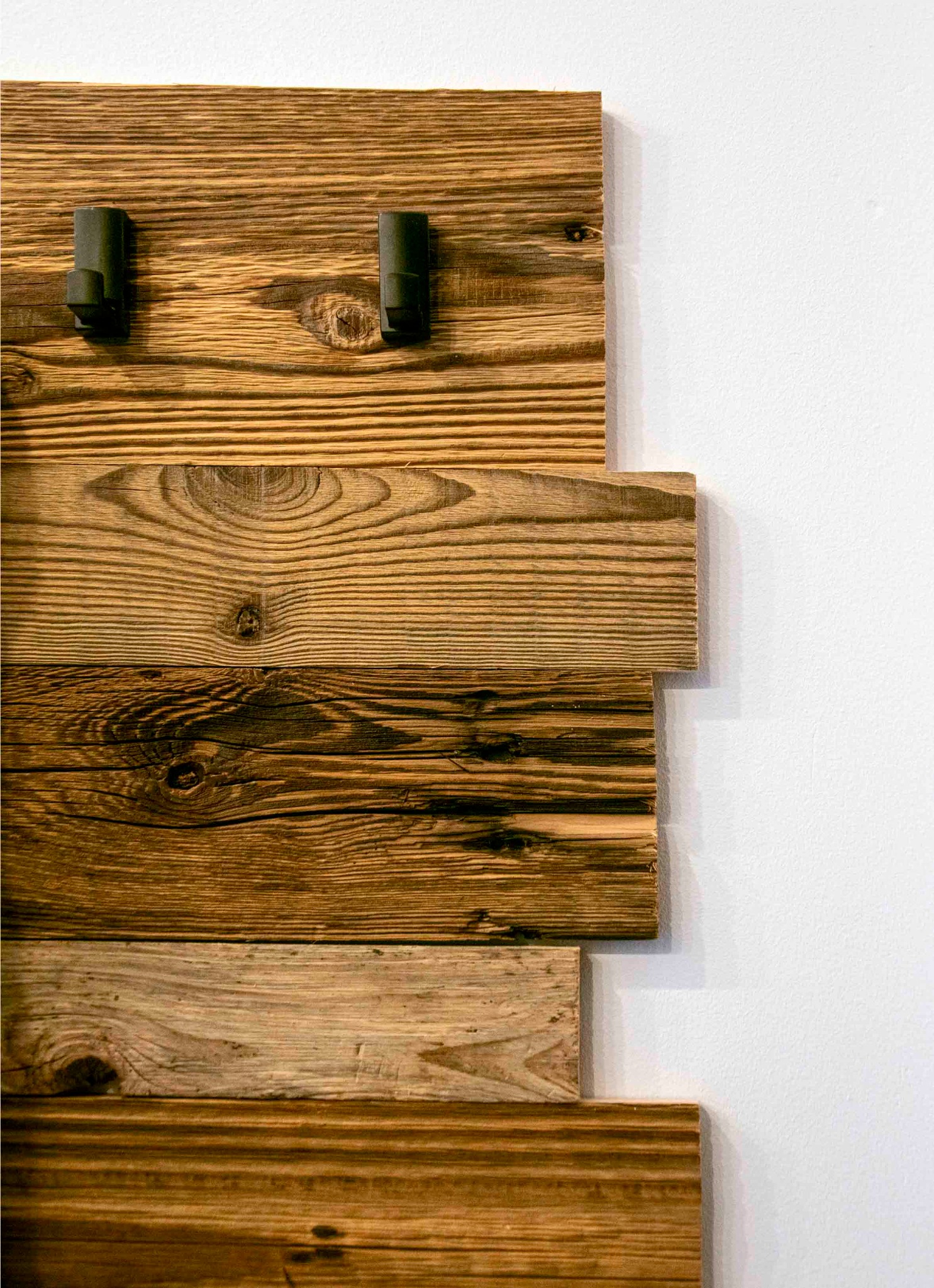 Dekoracyjna ściana z drewnianych desek o zróżnicowanej kolorystyce i fakturze, z dwoma czarnymi, metalowymi haczykami na górze, na tle białej ściany.