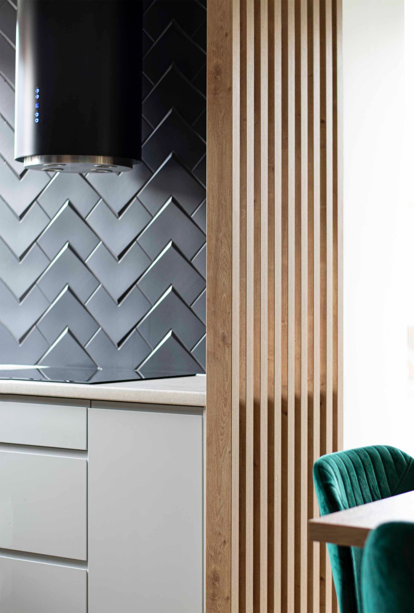 Częściowy widok nowoczesnej kuchni z szarymi płytkami ułożonymi w jodełkę, czarnym okapem, jasnymi szafkami i drewnianą ścianką działową, oraz zielonymi krzesłami w tle.