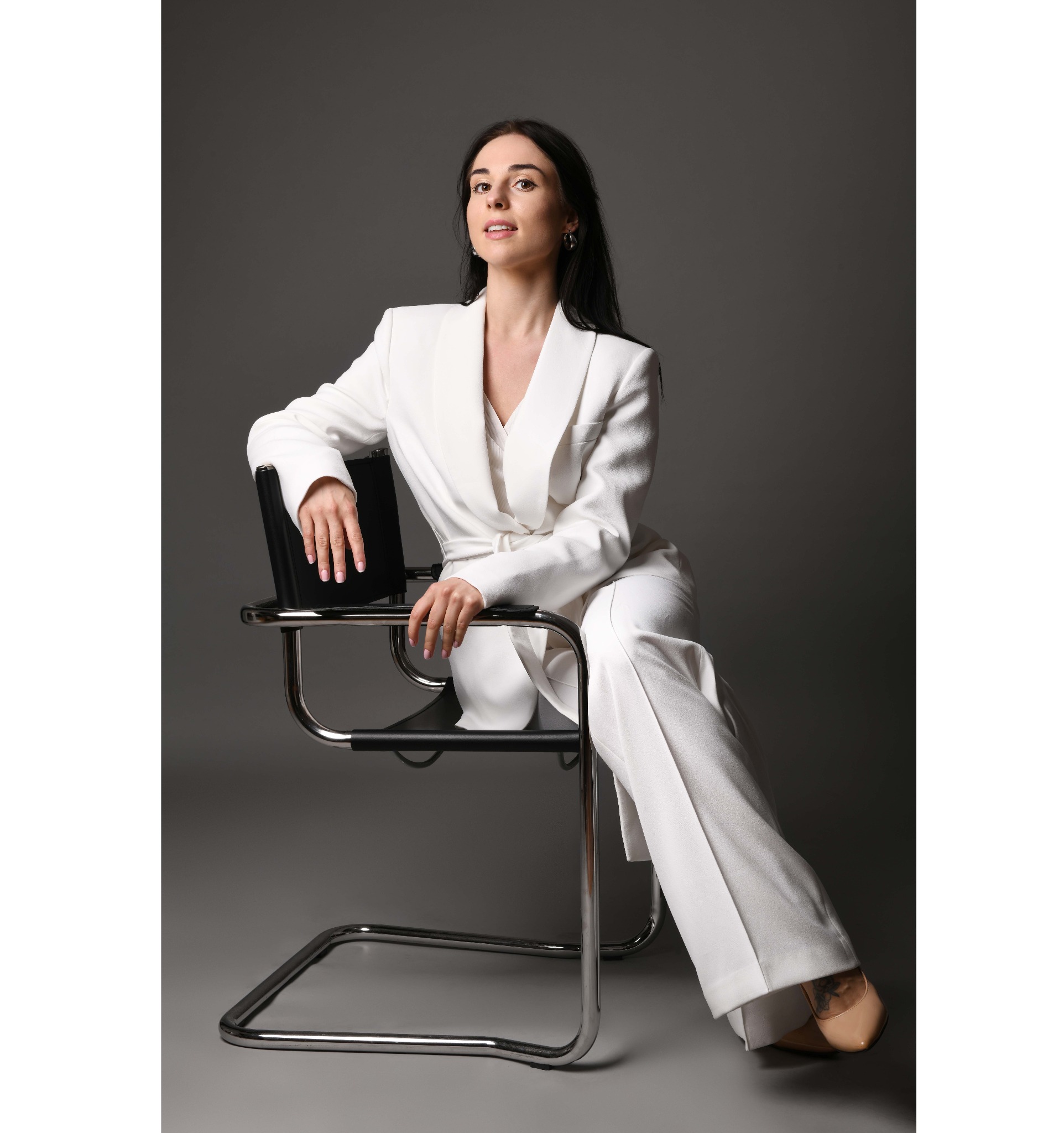 Kobieta w białym garniturze siedzi na krześle z chromowanymi nogami na tle szarego studia.