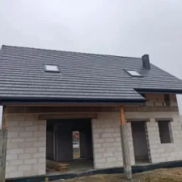 Dom w trakcie budowy z nowym, ciemnoszarym dachem z dachówki bitumicznej i oknami dachowymi, widoczne rynny, ściany z bloczków betonowych.