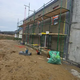 Budynek w trakcie ocieplania elewacji, widoczne rusztowania, styropian oraz narzędzia budowlane i worek na odpady.