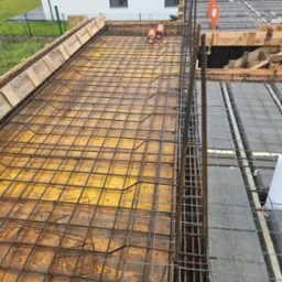 Zbrojenie stropu na deskowaniu z widocznymi elementami instalacji. Stalowe pręty tworzą siatkę, przygotowane do zalania betonem. W tle budynek w trakcie budowy.