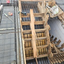 WOJCIECH KROPIDŁOWSKI - Zbrojenie stropu z drewnianym szalunkiem, widoczne pręty zbrojeniowe i elementy konstrukcji. Na pierwszym planie fragment buta pracownika.