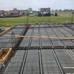 WOJCIECH KROPIDŁOWSKI - Zbrojenie stropu Teriva z pustakami szalunkowymi, widoczne pręty zbrojeniowe i pianka montażowa. W tle domy jednorodzinne i pochmurne niebo.